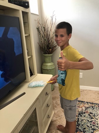 chores for 6-year-olds a boy dusting a tv stand while smiling and giving a thumbs up sign to the camera