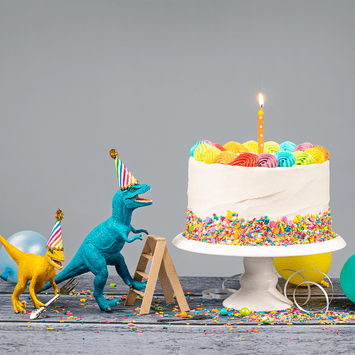 twins' birthday dinosaur figures wearing birthday hats, balloons, a white birthday cake with a candle and confetti on a table