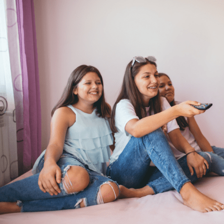 Three girls sitting on a bed watching TV The middle girl is sitting with her right leg bent and elbow resting on it while pointing the remote control at the TV. All there girls are smiling 