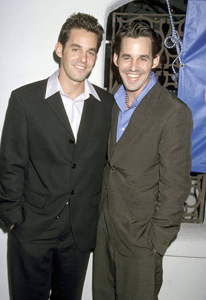 celebrity twin 2 men smiling at a camera in dark suits, 1 wearing a blue shirt, the other a white shirt.