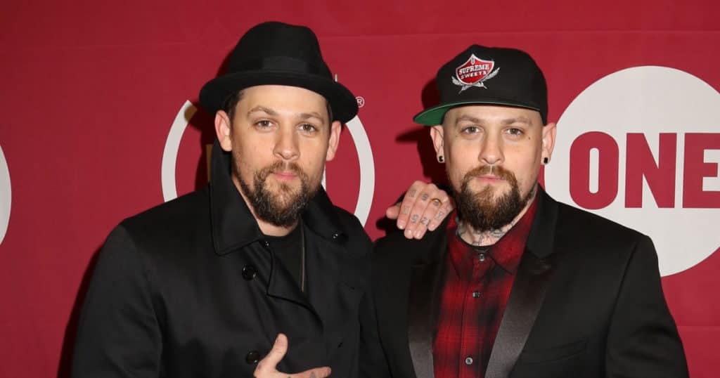 celebrity twins identical twin men posing, 1 with his hand on the others shoulder, each wearing a hat and black clothing.