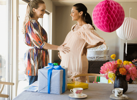 maternity baby shower dress woman holding baby bump while another woman rests her hand on her belly, they are in a decorated room for a baby shower