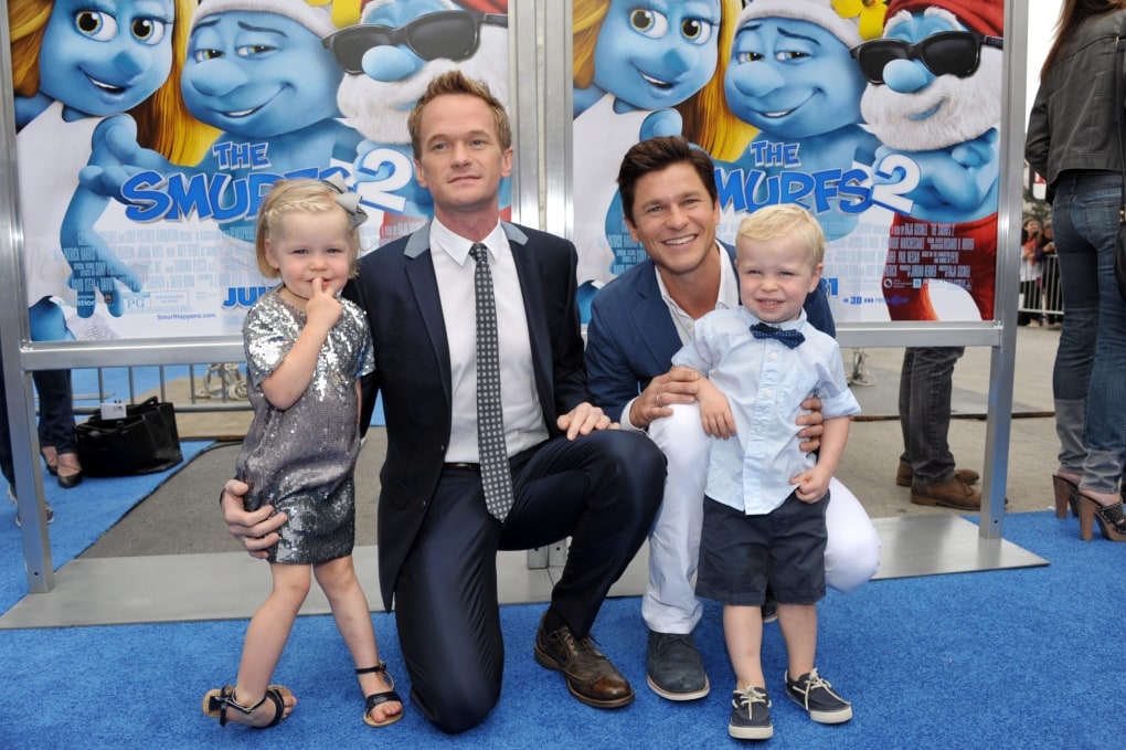 celebrities with twins Neil Patrick Harris and David Burtka posing with twin boy and girl on a blue carpet in front of a Smurfs movie poster