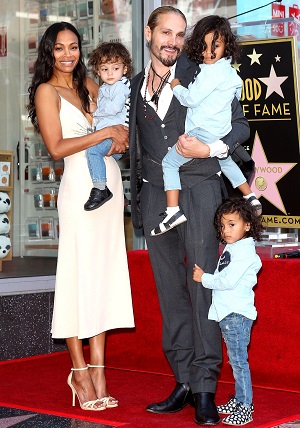 celebrities with twins Zeo Sldana and her husband each holding one small boy and another small boy holding the mans pants, all of them looking towards the camera