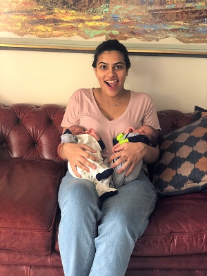 A new mom holding her infant twins, sitting on a brown couch