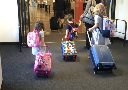 different parenting a woman holing a little boys hand, a little girl and an older girl, all pulling suitcases and wearing backpacks in an airport