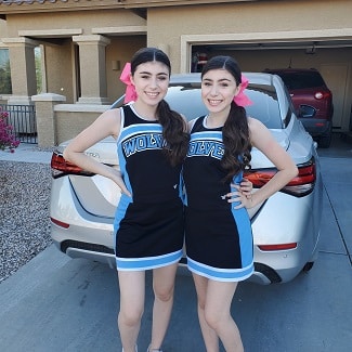 identical twin girls in cheer uniforms with bows posing in a driveway