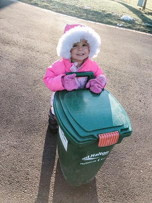 A toddler in a big pink coat pushing a trash can outside in a street