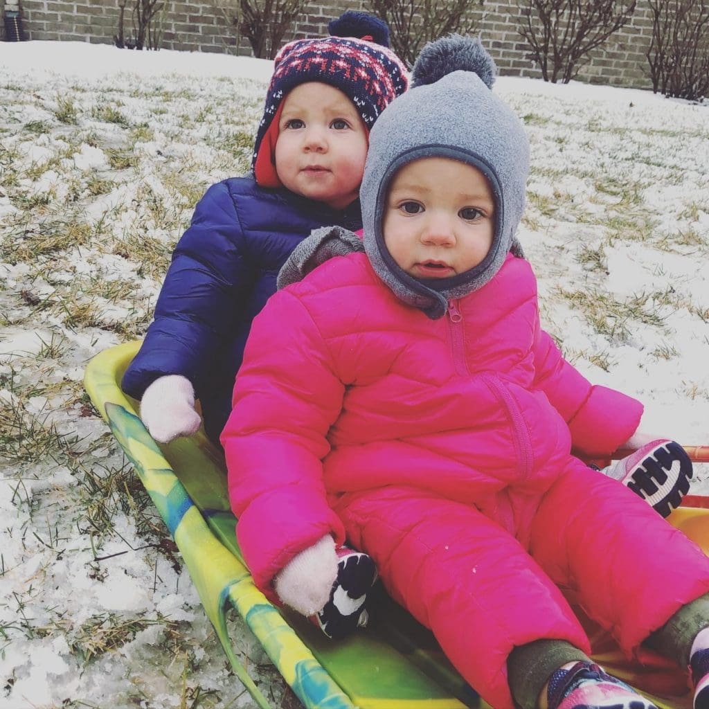 15-month-old identical twin girls on a sled in snow with snowsuits, hats, and mittens on.