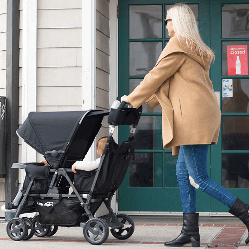 a woman pushing a tandem stroller with two kids on a sidewalk