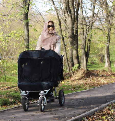 Woman in pink headscarf pushing black stroller along a paved trail in the woods