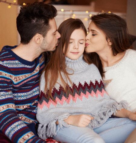 child with eyes closed wearing a sweater being kissed on the left cheek by mom with dark hair wearing a white sweater and right cheek by dad wearing a blue, magenta and white stripped sweater