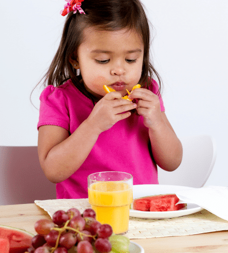 toddler girl sitting at a table eating an orange with melon and juice