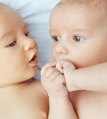 infant twins next to each other, 1 looking at the others' hands