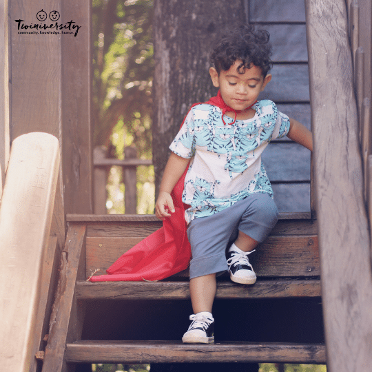 a boy with dark curly hair, a white and blue shirt and grey pants wearing a red cape walks down steps outside