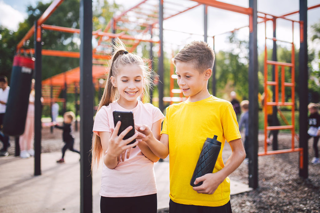 Preteen girl standing at the playground holding a cell phone smiling sharing the content with her twin brother who is does not have a phone but enjoys what he is seeing