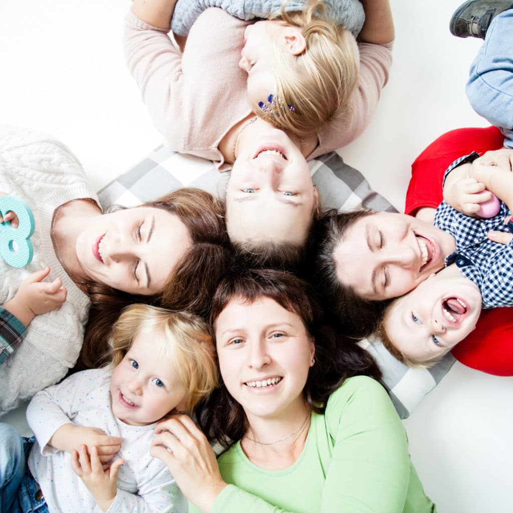 Group of 4 moms lying on their backs, heads together looking up, smiling and laughing as they play, tickle and snuggle their toddlers