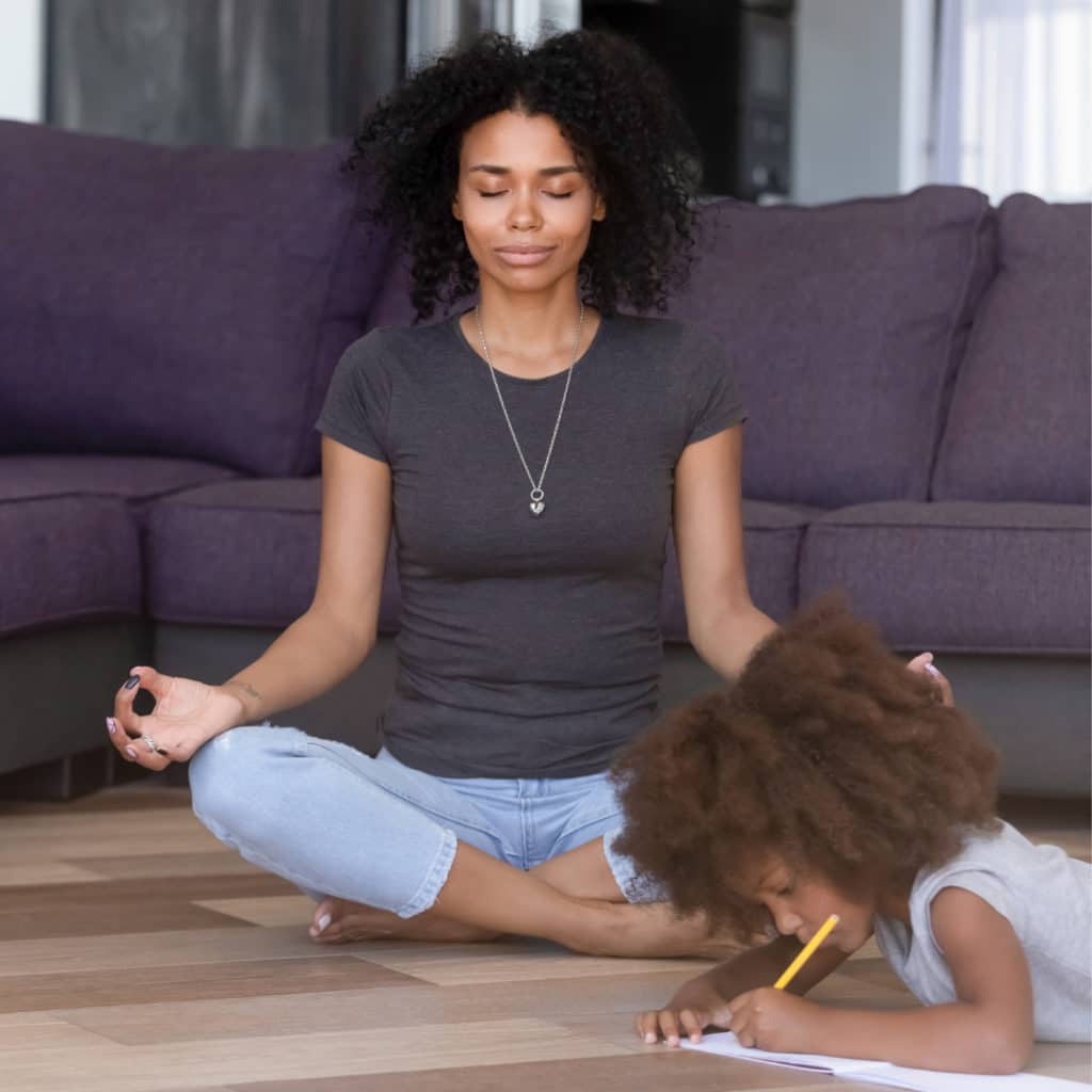 Mother sitting cross-legged on the ground of her living room eyes closed wrists resting on her knees, palms out meditating while her young daughter colors in note pad on the floor next to her