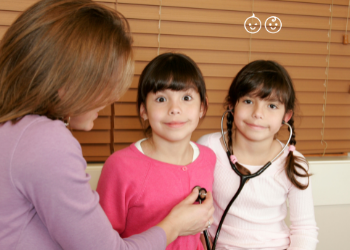 identical twin girls smiling at a camera with one twin listening to the others heartbeat with a stethescope