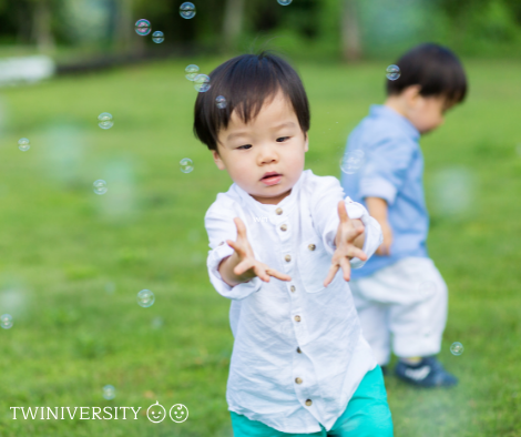 two dark-haired boys outside in the grass. 1 wearing a white shirt and chasing bubbles and the other wearing a blue shirt out of focus and looking at the grass