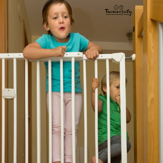 a young child standing at a baby gate with a blue shirt and white pants. A smaller boy in a green shirt and grey pants crouching beside her