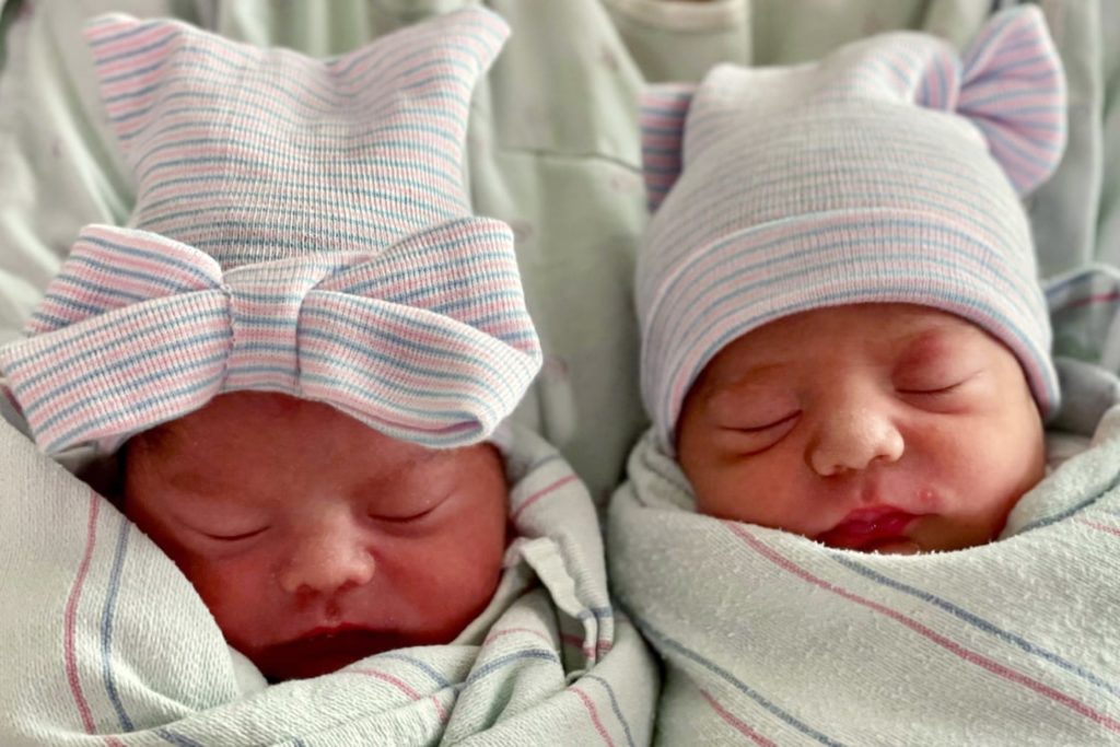 newborn baby twins born in separate years wrapped in blankets with pink and blue striped hats with bows