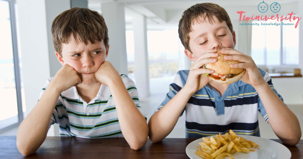 twin boys sitting next to each other with elbows on a table. 1 twin is eating a hamburger with french fries and the other is resting his face on his hands and scowling at the other. 