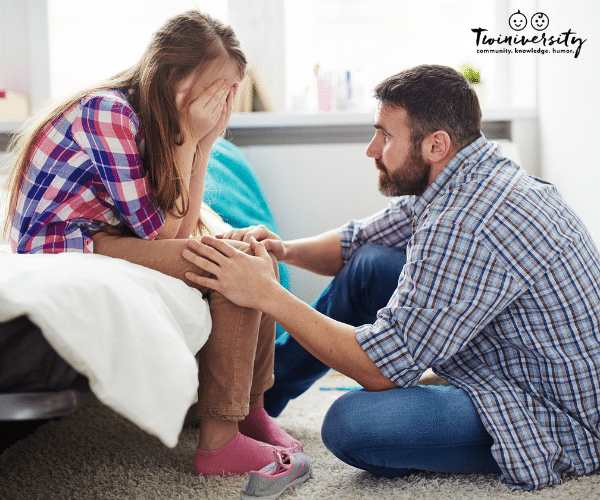 young girl sitting on bed with head in hands while adult male caregiver offers support with hands on her knees