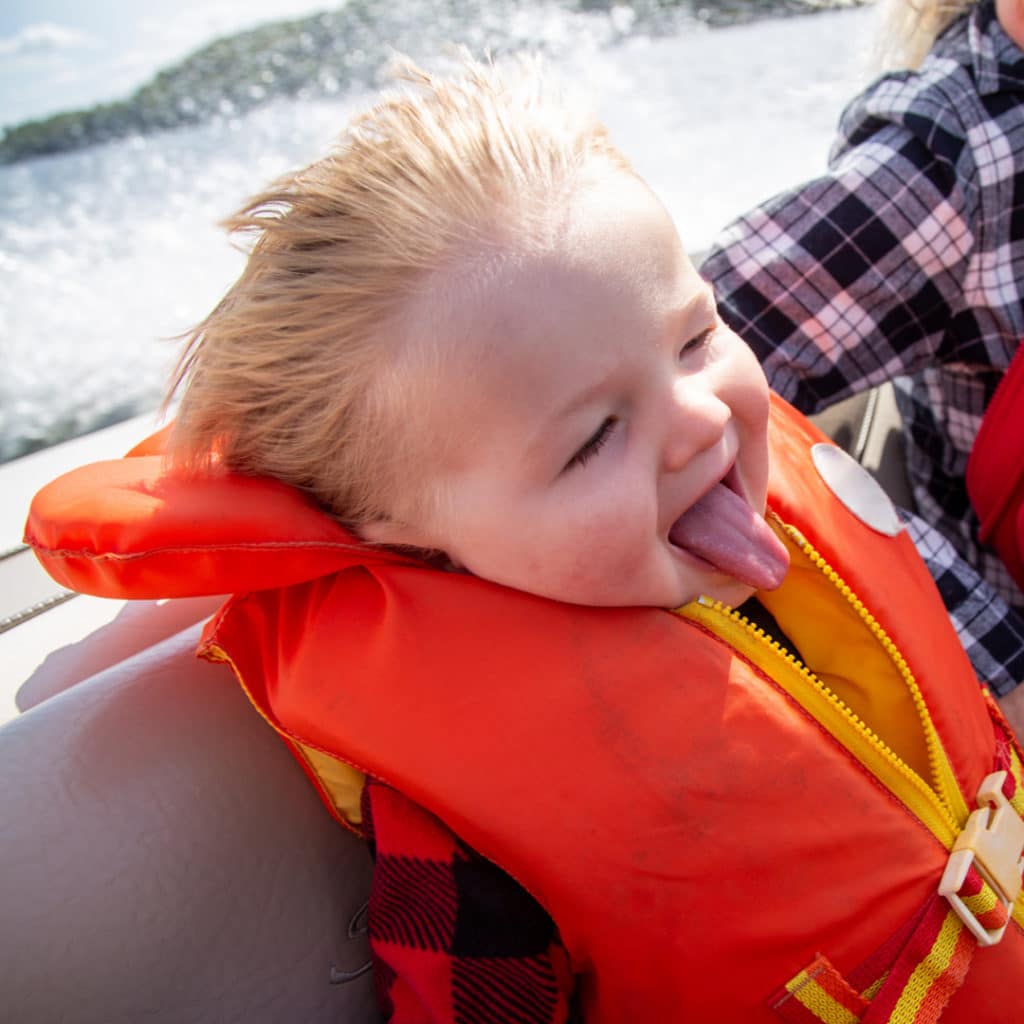 Toddler riding in a boat wearing an orange life jacket wind in his hair and tough sticking out in delight
