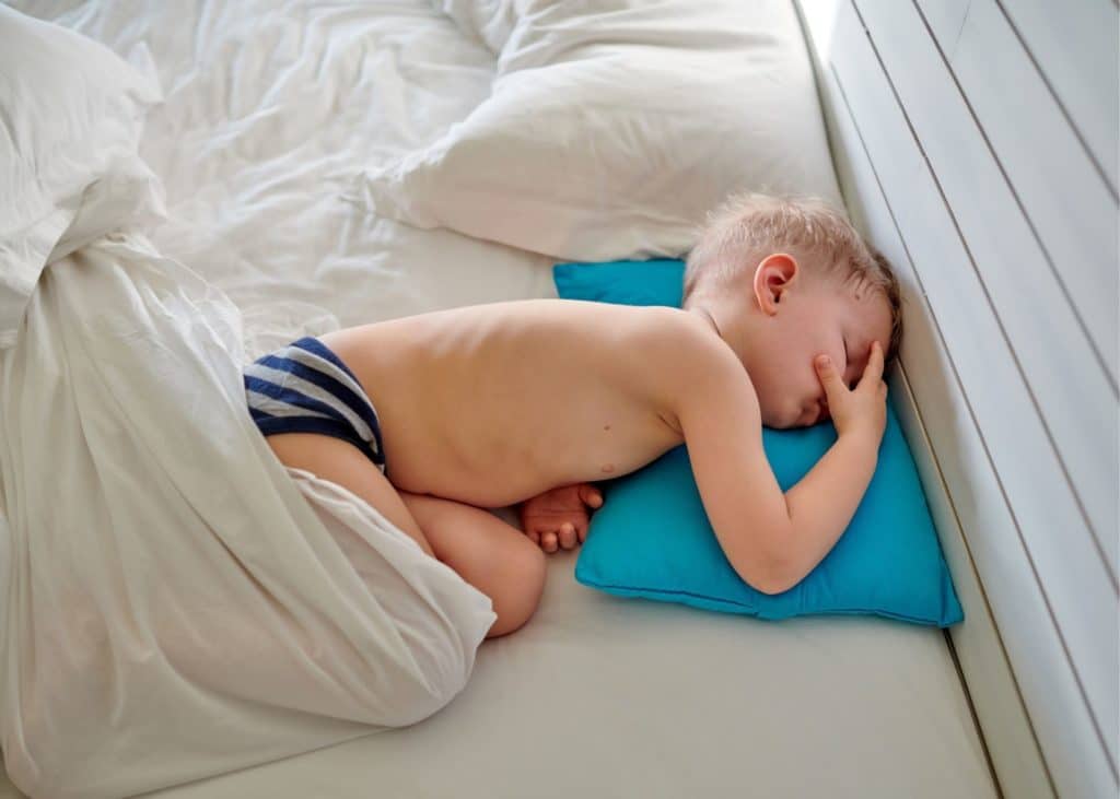 Toddler boy sleeping on a blue pillow wearing dark blue and gray stripped underwear. Hand is infant of his face and he seems to be sleeping in his parents bed made of white bedding