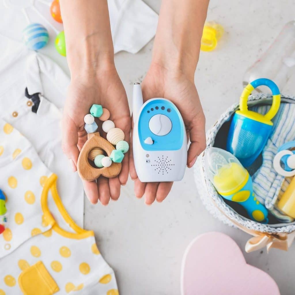 Female hands, palms up holding a wooden baby toy in her righthand and a baby monitor in the left hand. Background is full of other baby items, including onesies, pacifier and toys.