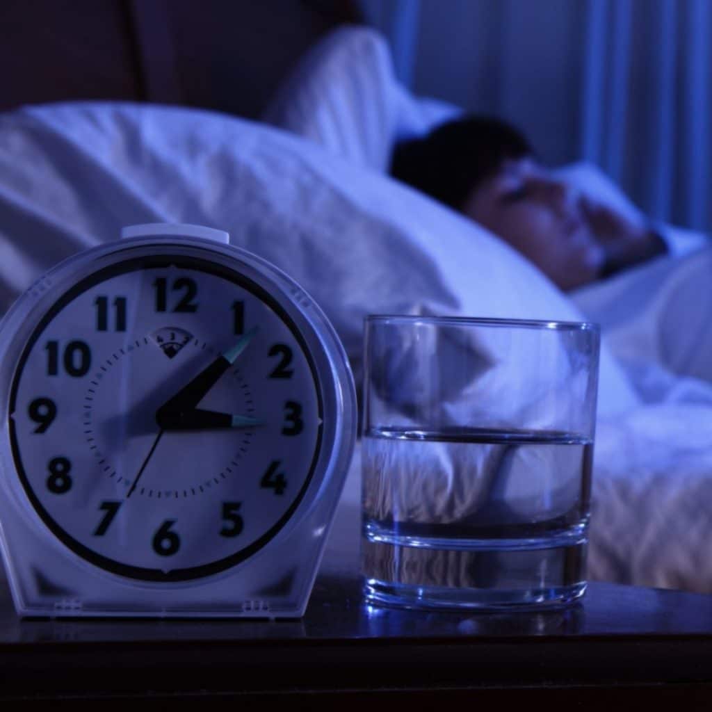 Young child sleeping in the background with a clock set at 3:07 am and a glass half full of water sitting on a table in the front of the image
