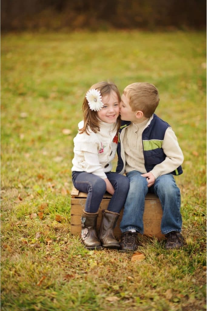 a young boy kissing a young girl on the cheek while sitting on a box in grass
