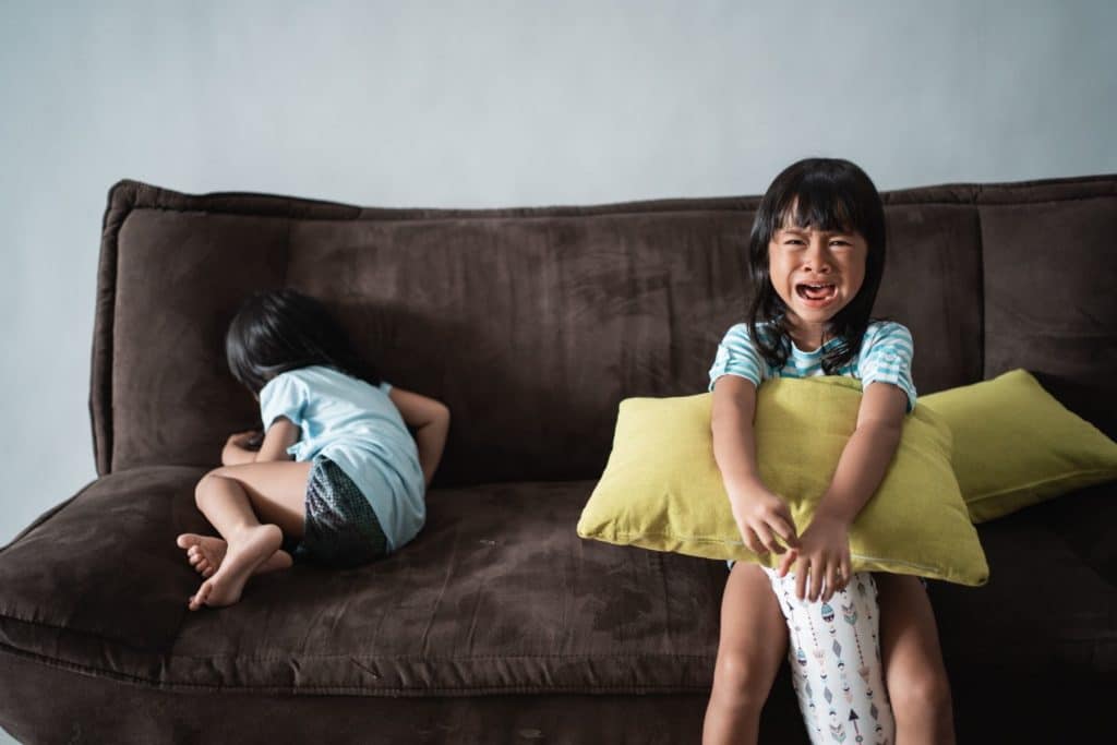 a young girl holding a pillow, crying on a couch and another young child curled up against the corner of the same couch. 