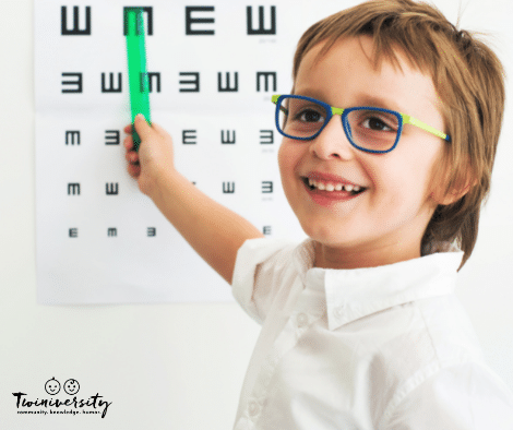 young boy wearing glasses smiling in front of eye exam chart 