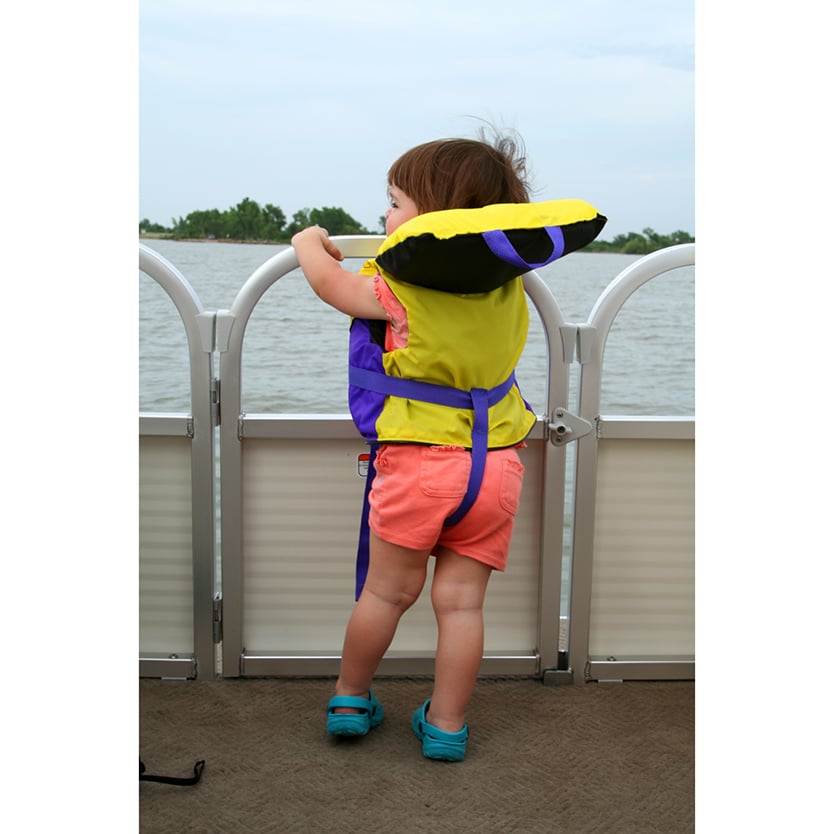 Toddler wearing a yellow and purple life jacket standing water side holding on to the railing