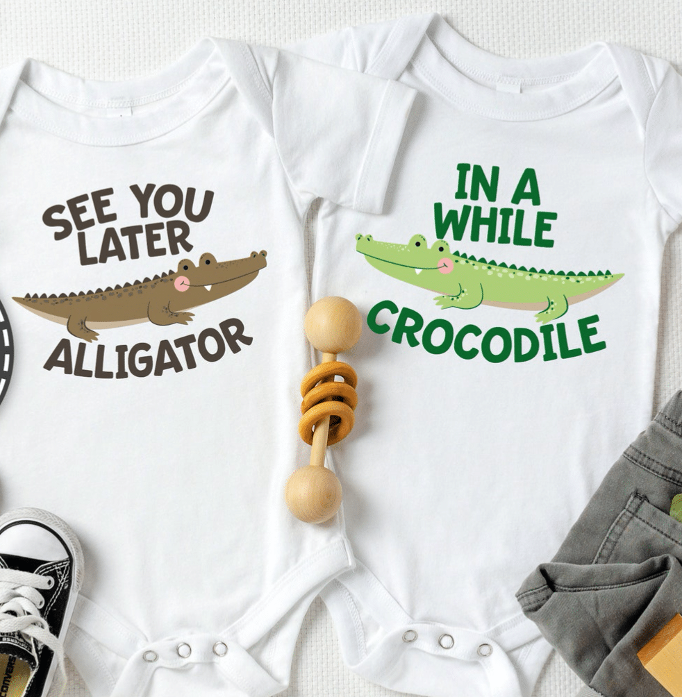 White onesies situated side by side, left reading "See you later Alligator" with a graphic of a brown crocodile, and the right reading "In a While Crocodile" with a graphic of a green alligator.  Onesies are surrounded by a black sneaker, a rattle, and gray pants.