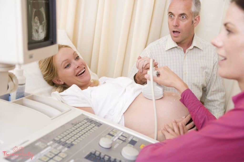 A middle-aged couple is having an ultrasound done. Mom is smiling and lying on a hospital bed, with the ultrasound wand on her belly. Dad is looking at the ultrasound screen with a shocked look on his face.