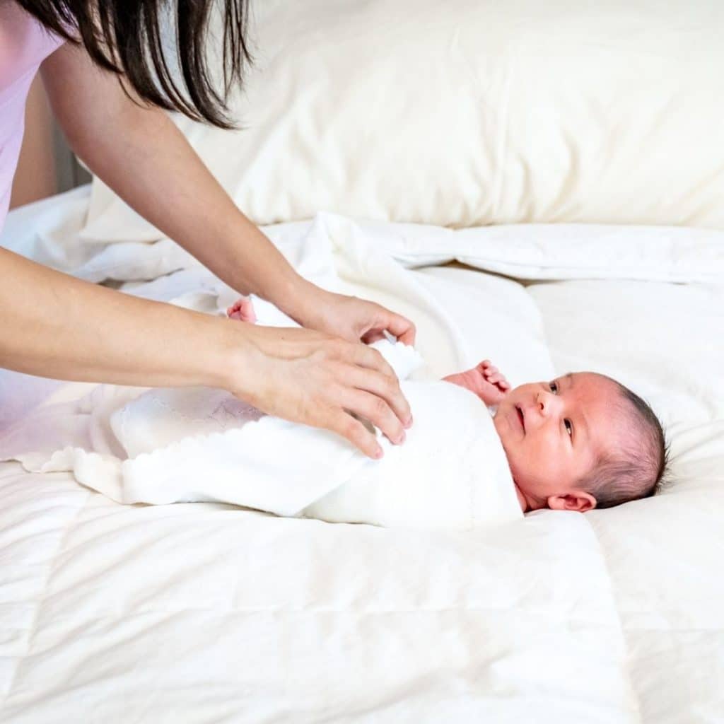 Newborn baby lying on a bed, looking up at their mom who is swaddling them in a white blanket.