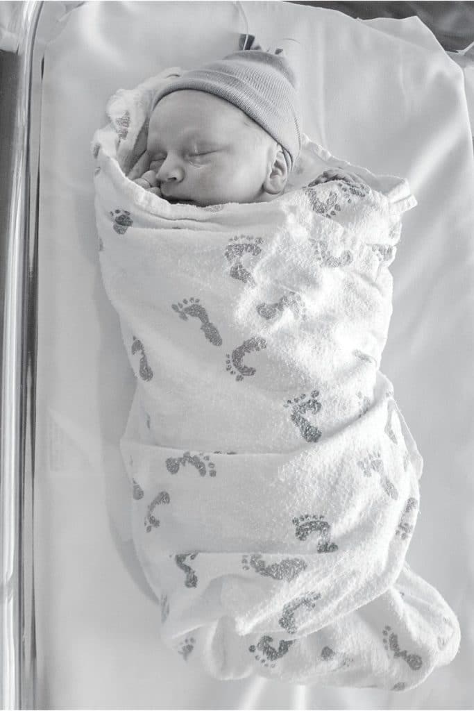 Newborn baby swaddled in a hospital blanket with a baby foot print on it, sleeping in a hospital bassinet.