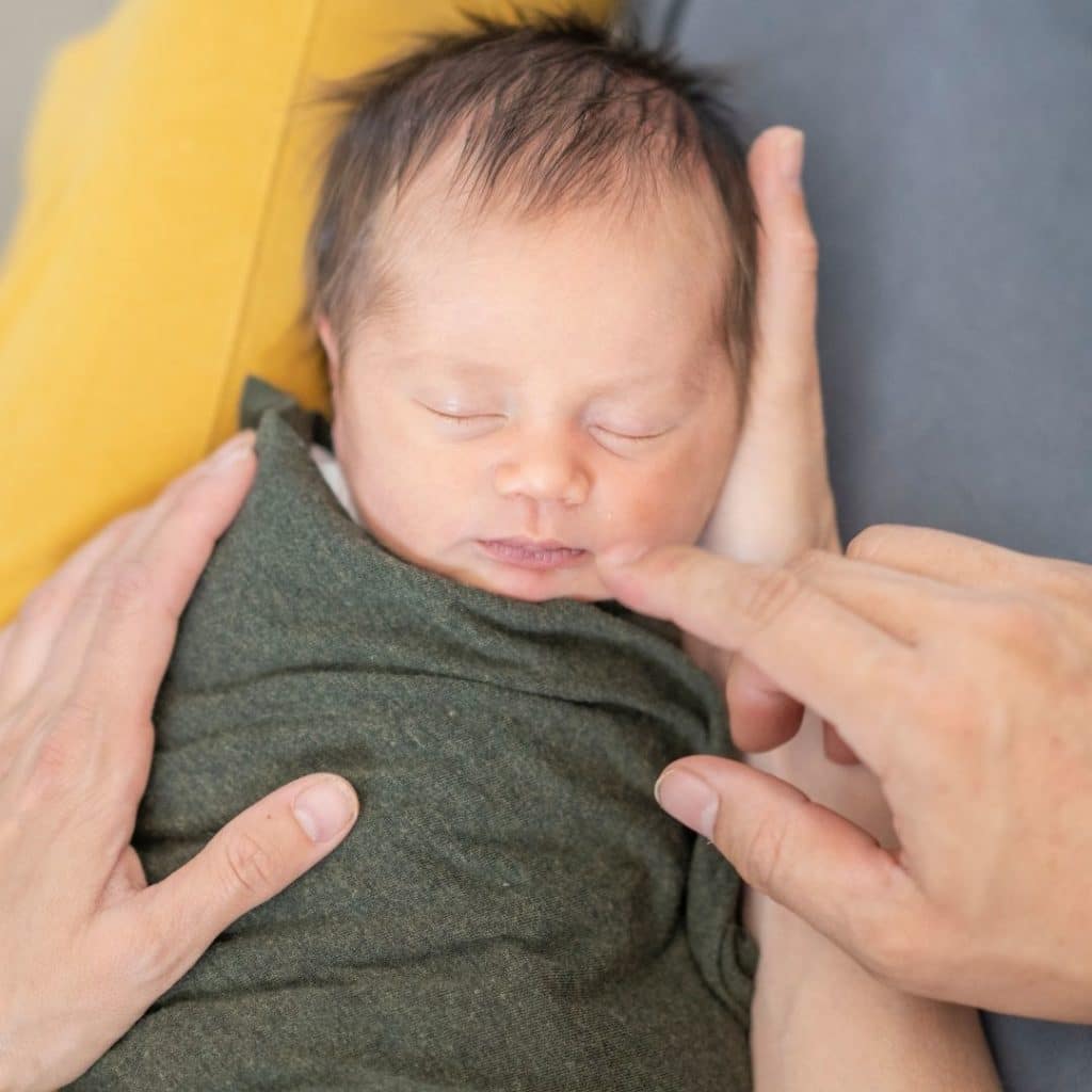 Newborn baby with lots of dark hair is swaddled in an army green cotton swaddle blanket while being held and admired by both mom and dad. 