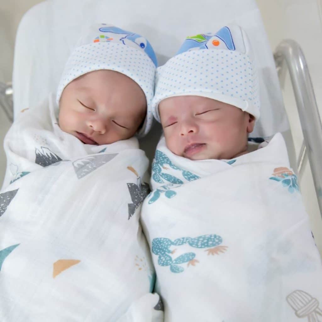 Newborn twins swaddled individually in muslin swaddle blanket, sleeping next to each other in a hospital bassinet.