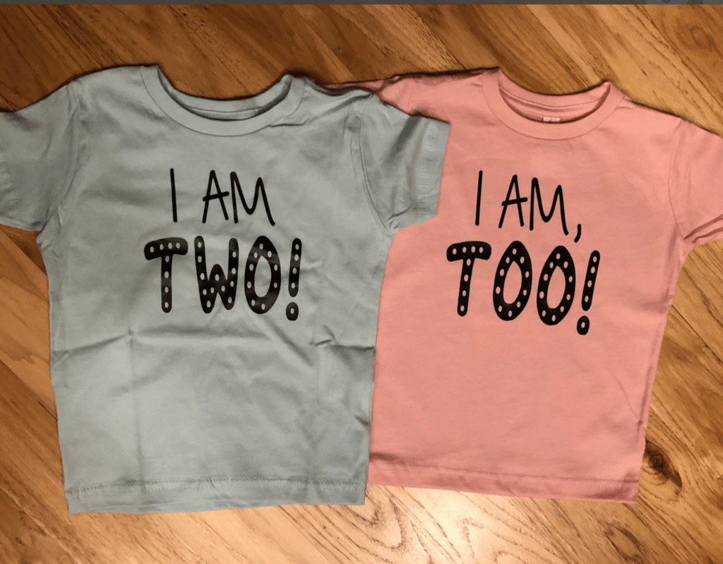 Tee shirts situated side by side; left side in light blue with black lettering reading "I am Two!'; right side in pink with black lettering reading "I am, too!"