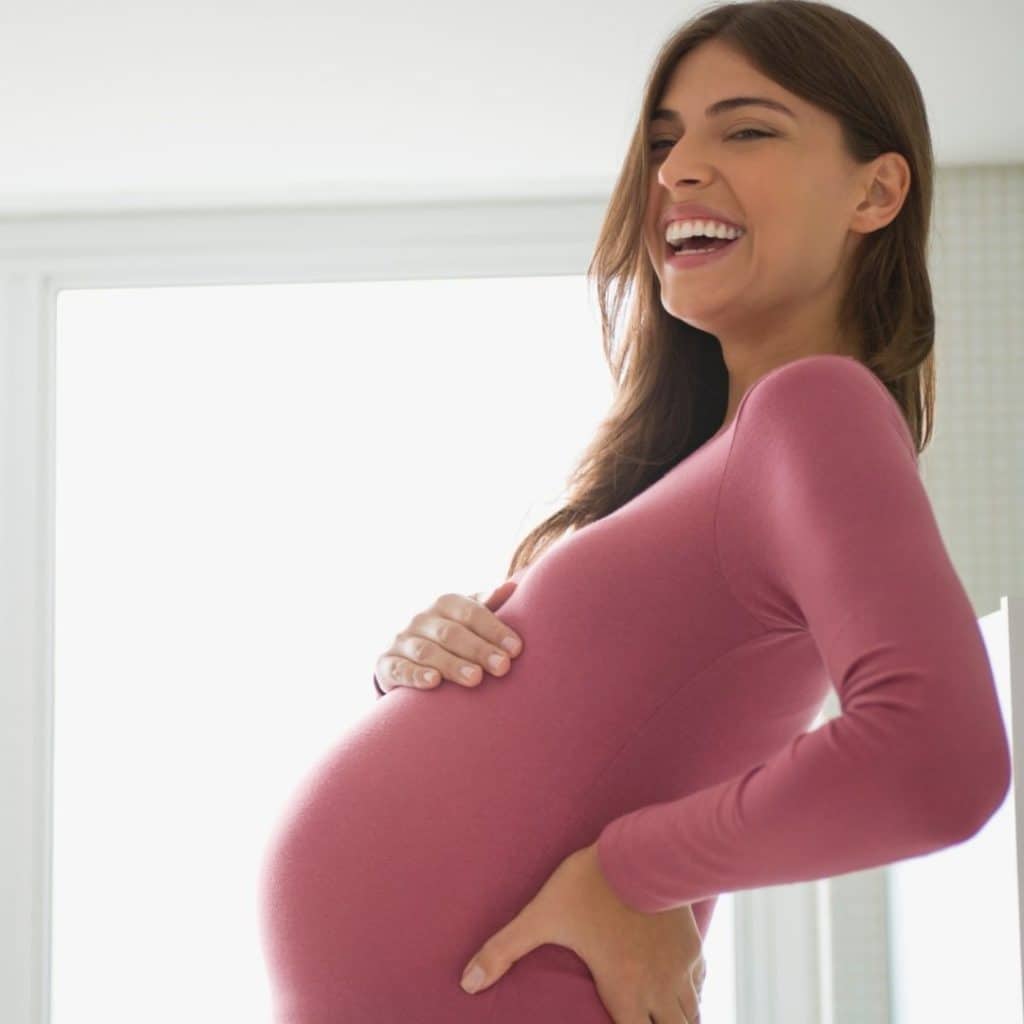 Pregnant woman wearing a mauve long-sleeve shirt standing in front of a window laughing and resting her left hand on her baby bump