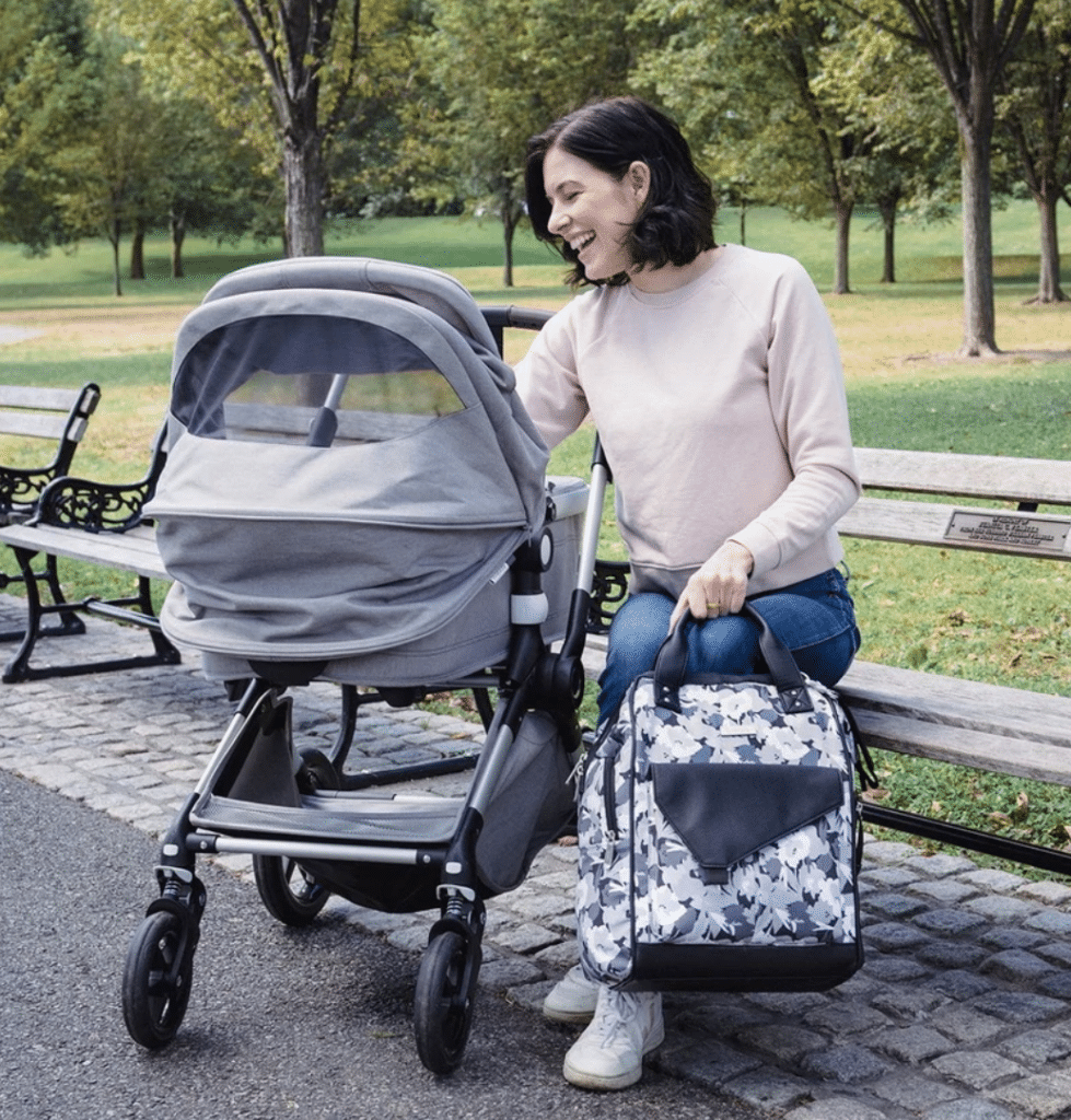 a woman sitting on a bench outside looking into a stroller facing away. She is holding a black, white, and grey floral print diaper bag.
