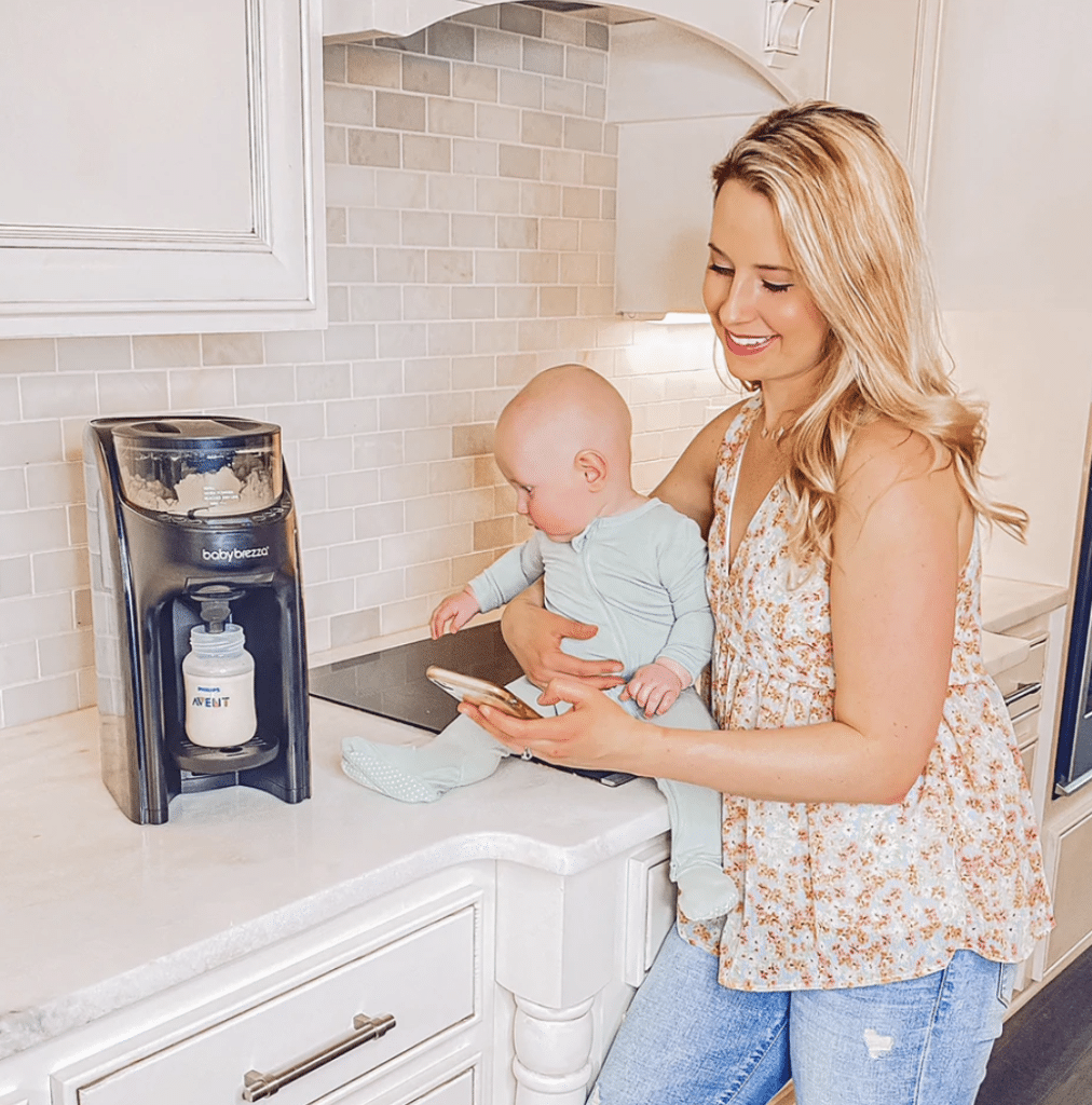 a blonde woman standing in a kitchen holding a baby with a green sleeper on in one hand and her phone in the other as her baby watches a formula maker dispense a bottle for twin feeding hacks