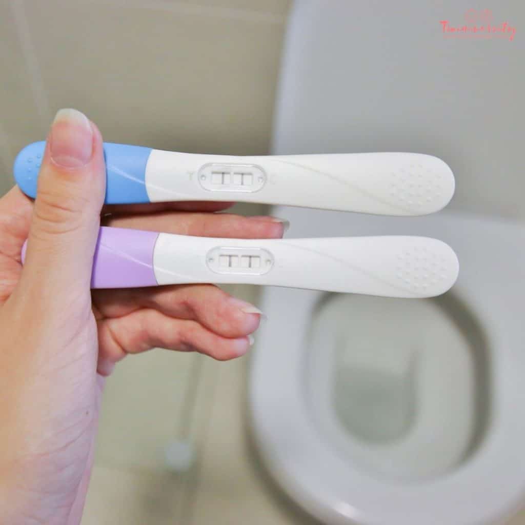 A woman is holding two pregnancy tests in her left hand over a bathroom toilet. One test is blue and one is pink, and both are showing a positive result