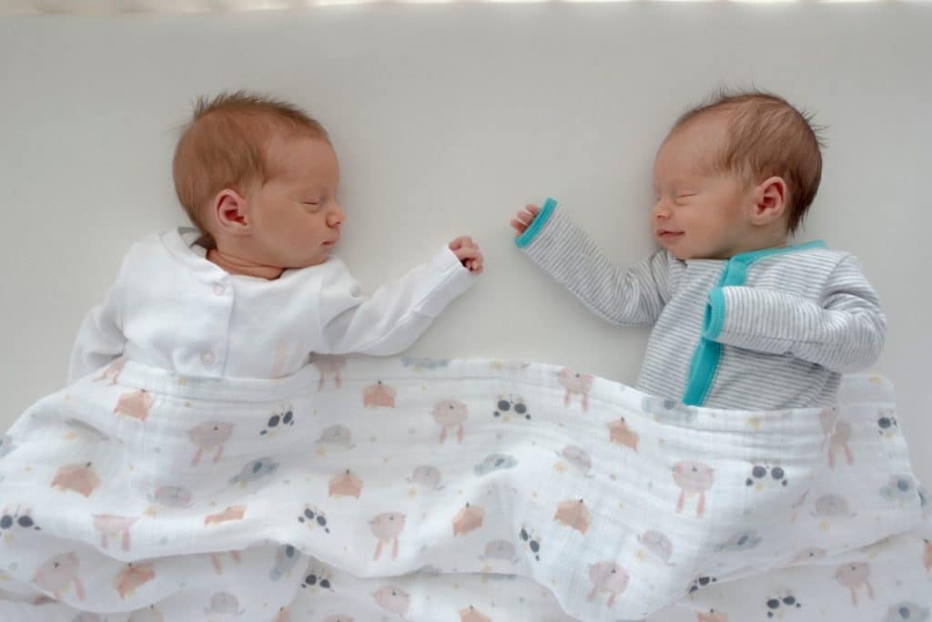 Twin babies sleeping next to each other, wearing long sleeve onesies with a swaddle draped over their legs. Baby on the right is smiling. Caption says 'if i don't need two of everything, what do i need for a twins baby registry?'