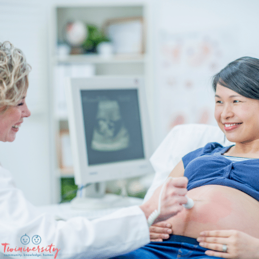 A female doctor has a Doppler on a pregnant womans belly with an ultrasound screen out of focus in the back.