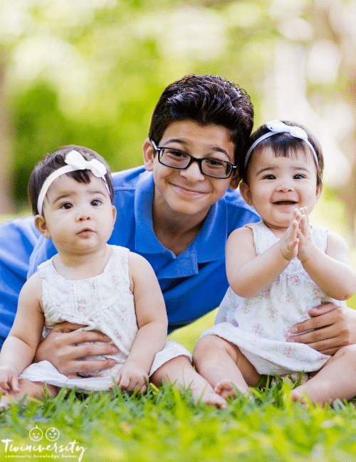 a young boy in glasses sitting with infant twin girls outside in grass